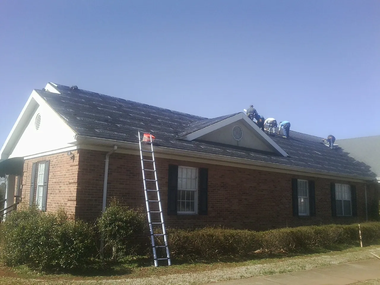 Crew tearing shingles off a brick ranch roof with a ladder set — Metal Roof Repair work by our crew in Donalds