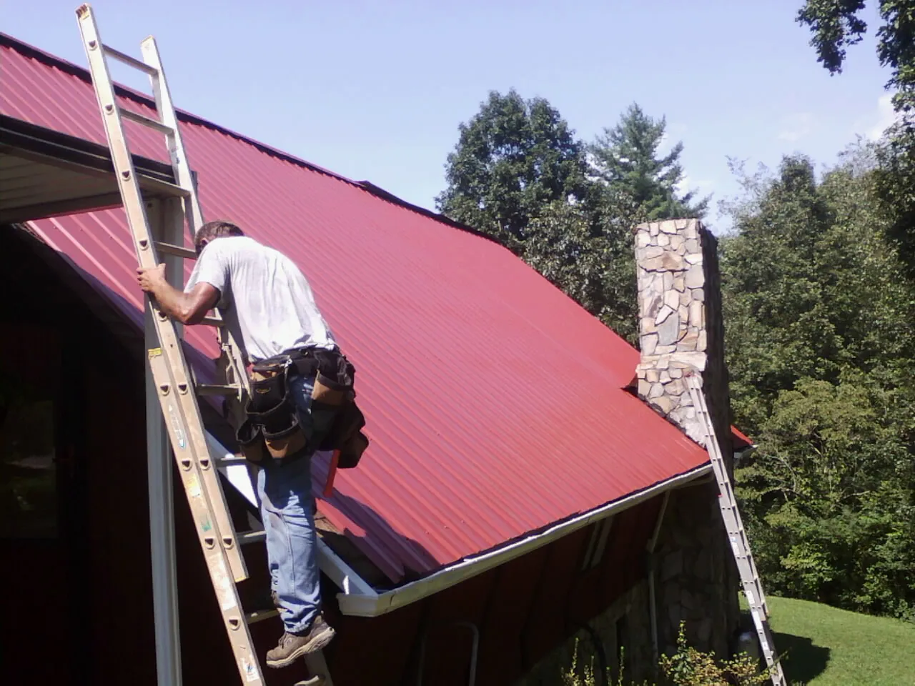 Roofer on a ladder beside a red metal roof and stone chimney — Soffit & Fascia Repair by Roofing Brothers in Donalds